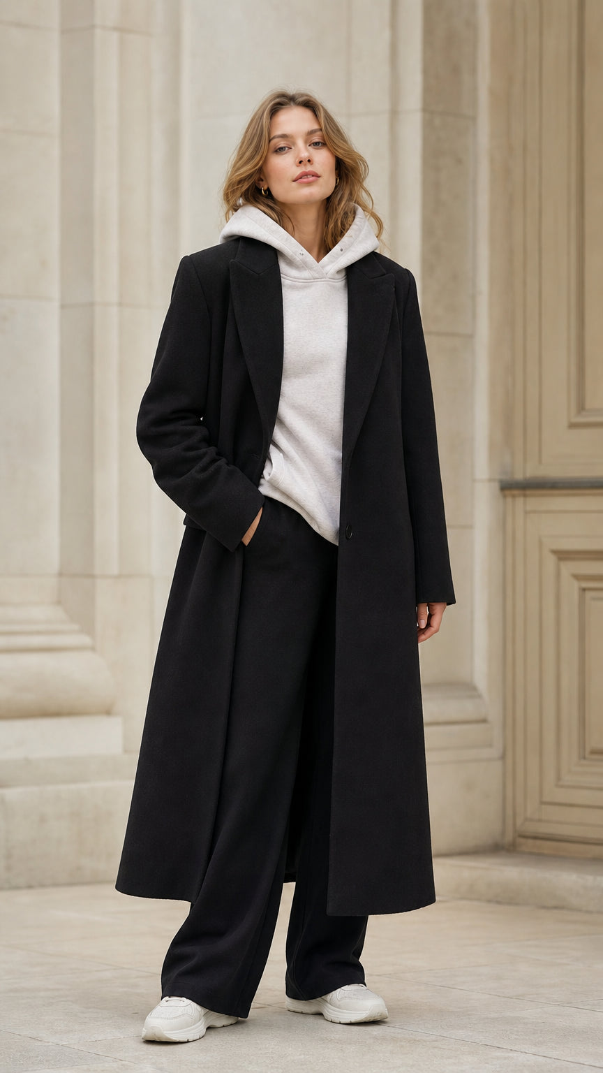 Front view of model wearing black tailored longline overcoat with white hoodie and wide pants
